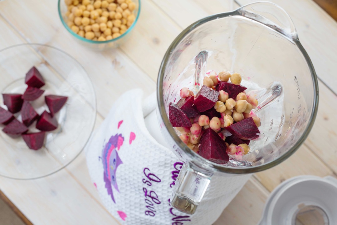 A glass blender jar with measurement markings filled with chopped beets and chickpeas on a kitchen counter
