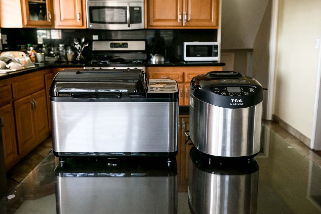 Two bread machines side by side on a kitchen counter, a wide horizontal-loaf model and a taller vertical-loaf model