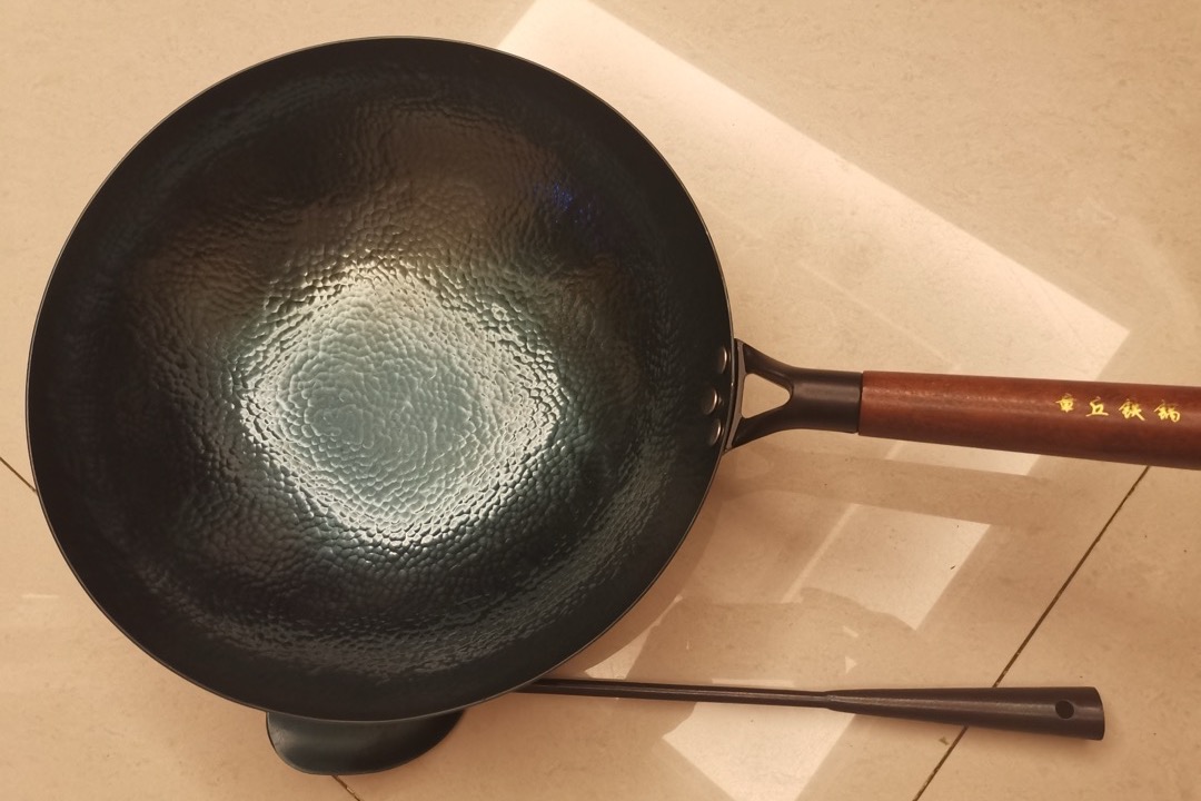 A black seasoned iron wok with a long wooden handle photographed from above on a pale surface