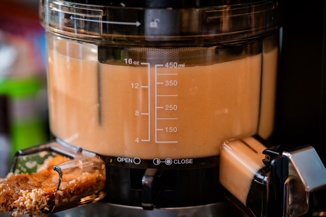 Close-up of a vertical cold press juicer chamber full of pale juice, with a measuring scale on the side and pulp at the outlet