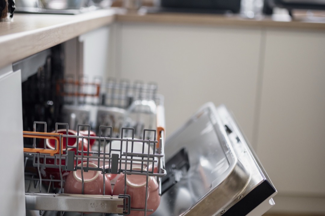 A built-in dishwasher with its door folded down and the upper rack pulled out, holding pink mugs, under a wood countertop
