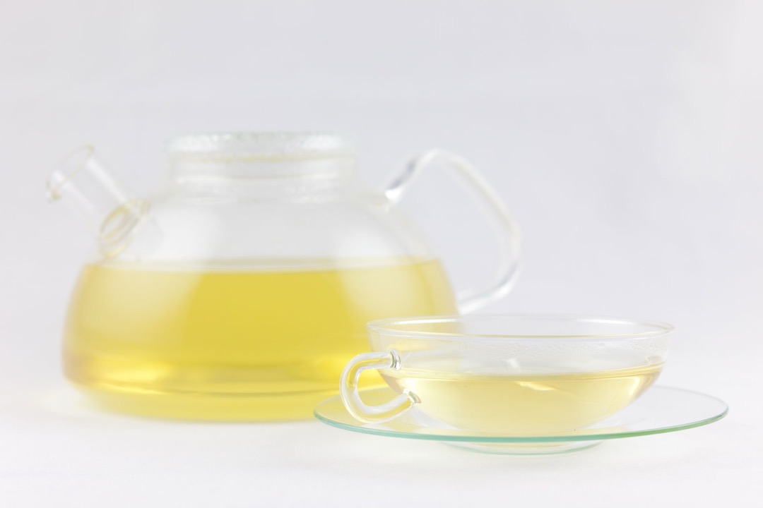 A clear glass teapot of pale brewed green tea beside a matching glass cup and saucer