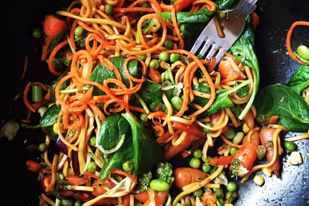 A stir-fry of noodles, carrots, spinach and peas cooking in a dark wok seen from above
