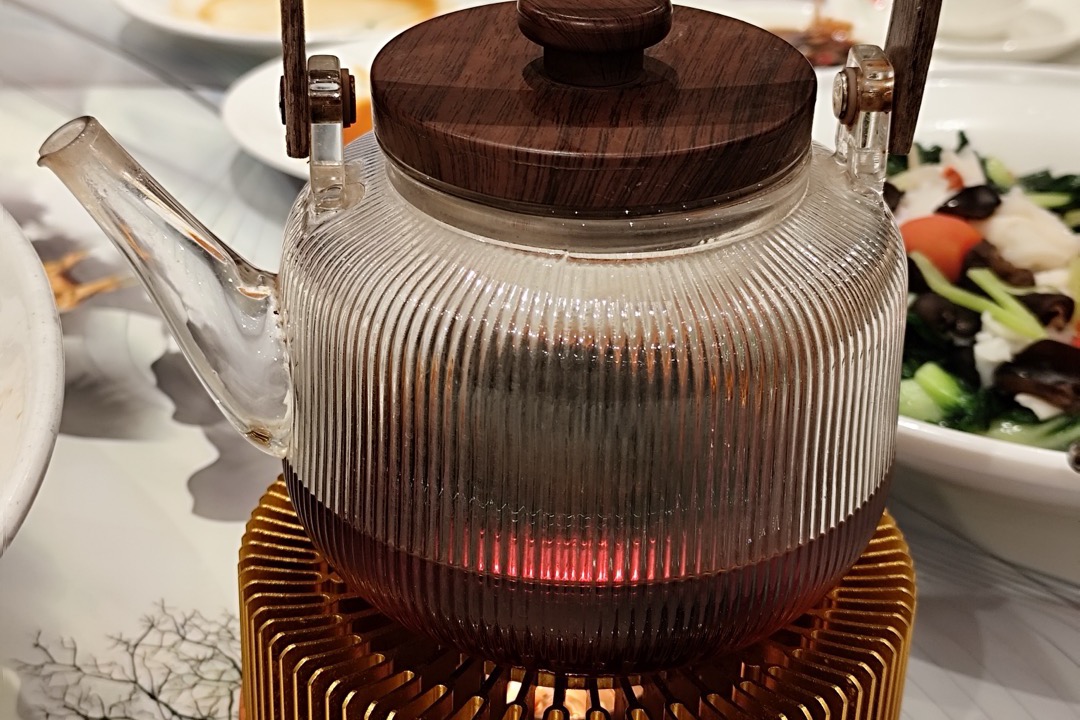 A ribbed borosilicate glass kettle with a dark wooden lid and handle warming on a brass stand
