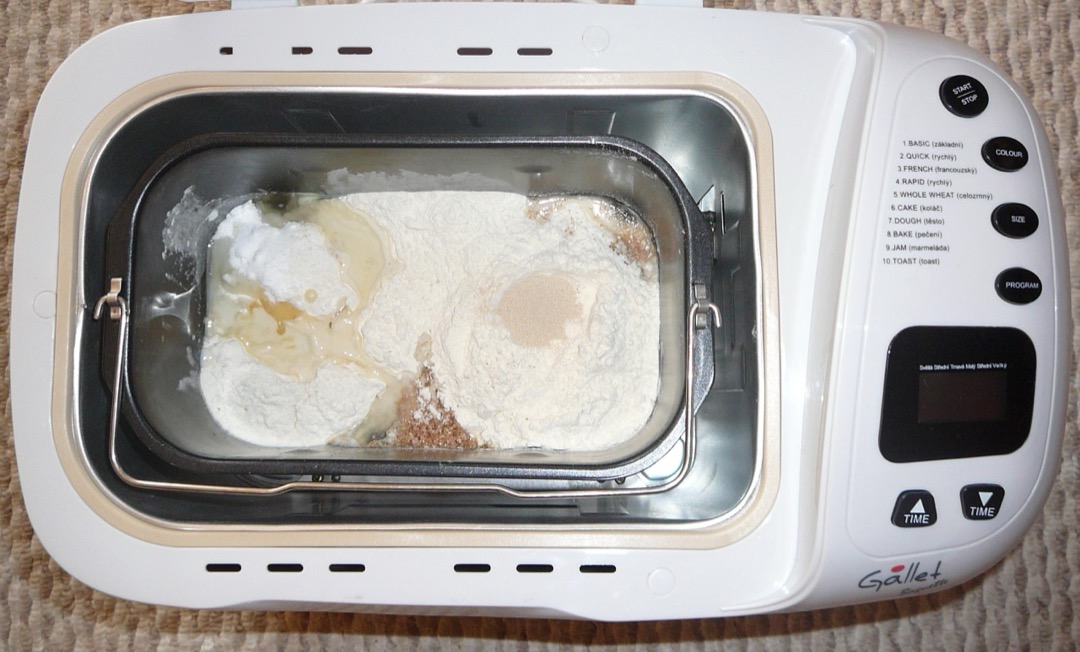 A bread machine on a kitchen counter beside a bag of flour, a bowl of gluten-free flour blend, and baking ingredients