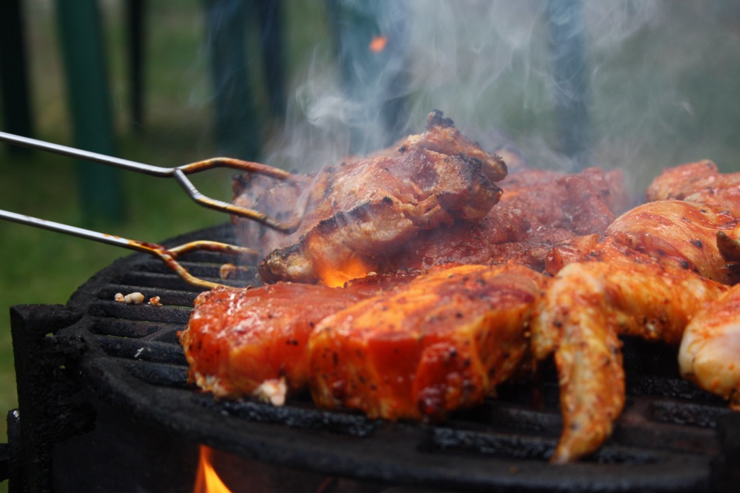 Long stainless steel grill tongs turning pork and chicken over the open flame of a charcoal grill