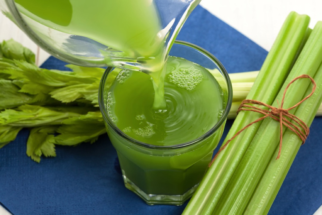 Green celery juice being poured from a glass jug into a tumbler beside a tied bundle of celery stalks on a blue cloth
