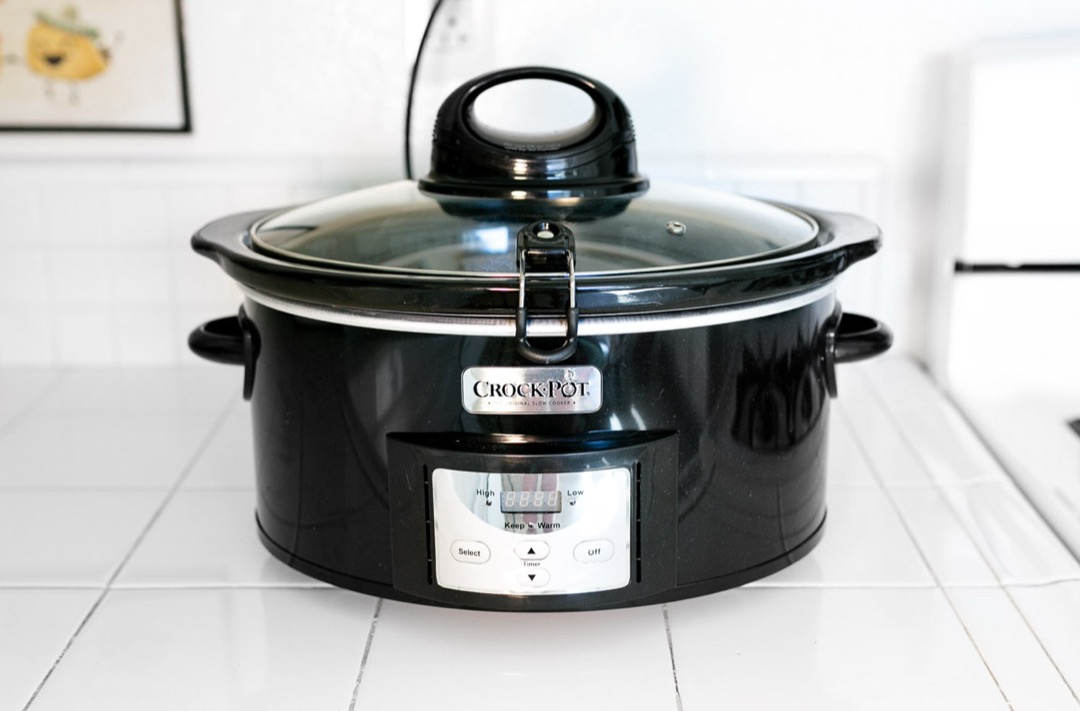 A black programmable slow cooker with a glass locking lid on a white tiled kitchen counter