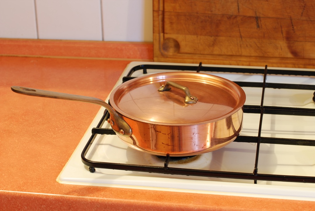 A lidded copper saute pan with straight sides sitting on a white gas stove