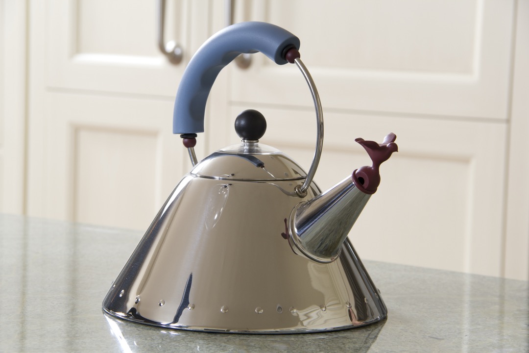 A mirror-polished stainless stovetop tea kettle with a blue handle and a bird-shaped spout whistle, standing on a stone counter