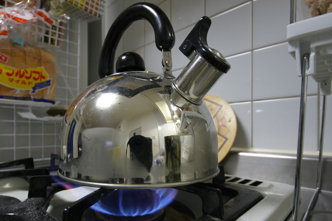 A stainless whistling tea kettle sitting on a lit blue gas flame on a black burner grate