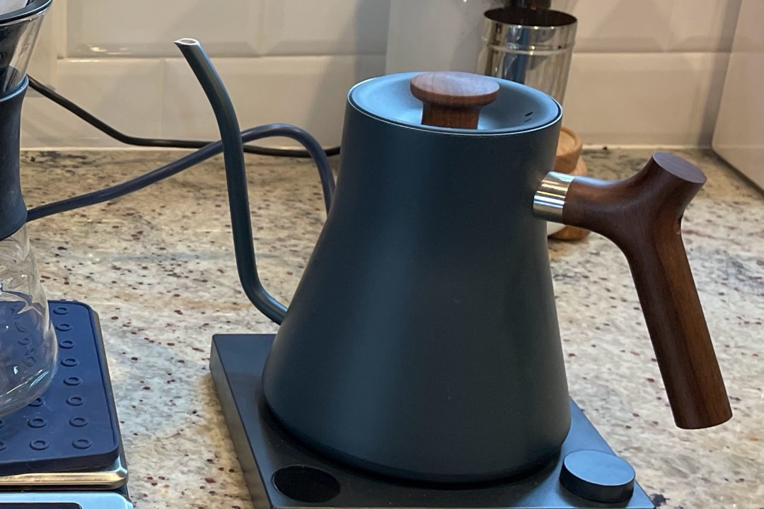 A matte black gooseneck electric kettle with a wooden handle sitting on its powered temperature-control base on a kitchen counter