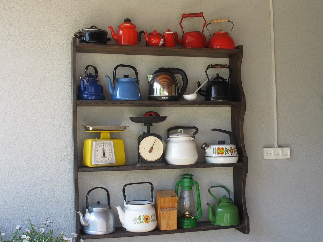 A wall shelf of enamel stovetop tea kettles in red, blue, white and green alongside a stainless electric kettle
