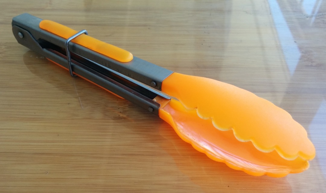 A silicone-tipped locking kitchen tong on a bamboo board, showing the pull-ring lock and scalloped gripping heads