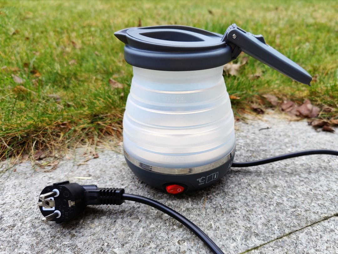 A collapsible silicone travel kettle with its folded body and lid open, plugged in on a stone step