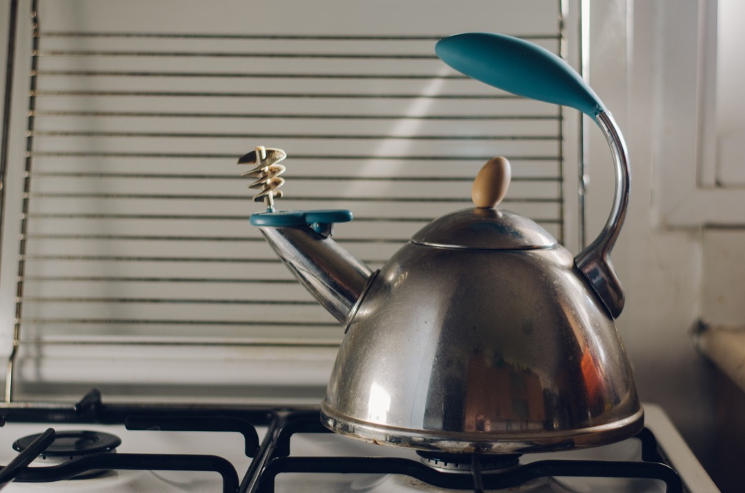 A stainless whistling tea kettle on a gas hob with its whistle cap flipped open, lit by warm window light