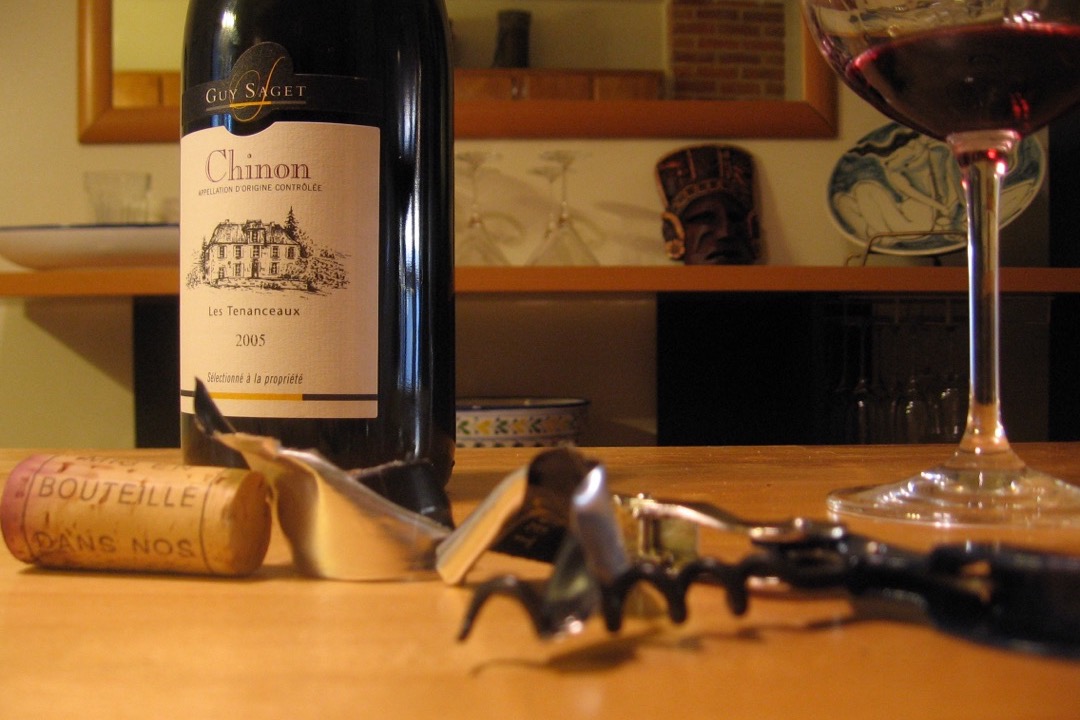 A waiter's corkscrew with a pulled cork and a strip of cut foil on a wooden table, in front of a bottle of Chinon red wine and a half-full glass