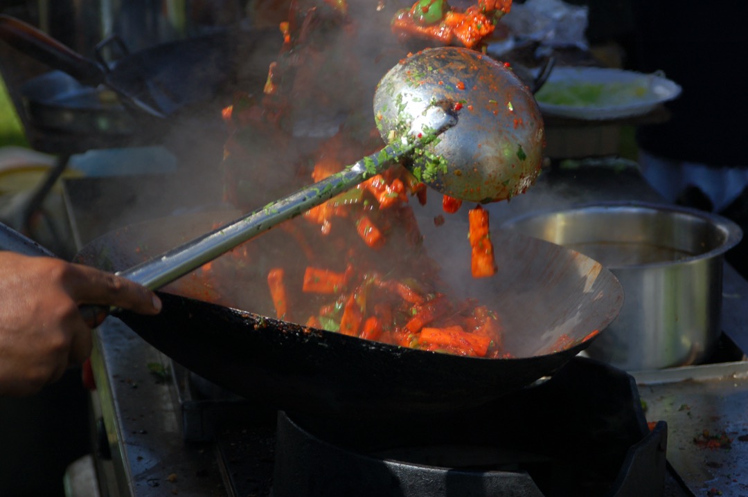 A carbon steel wok on a high-output burner with vegetables mid-toss and a metal wok ladle