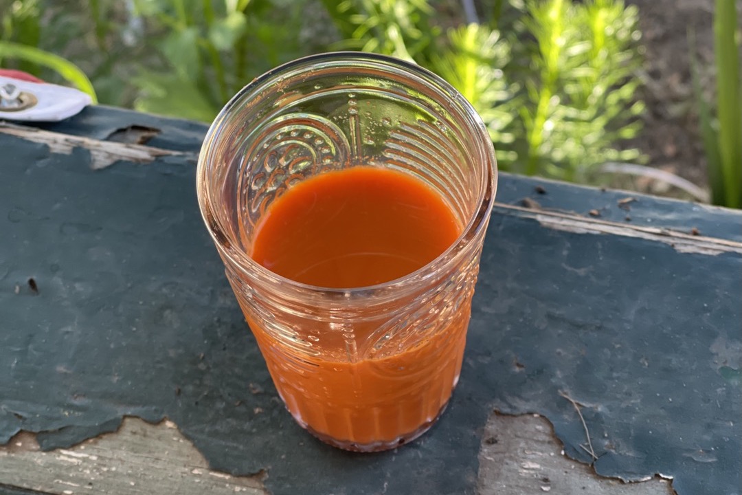 A ribbed glass tumbler of bright orange fresh carrot juice standing on a weathered green wooden bench outdoors
