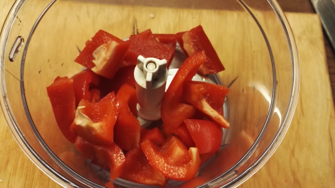 Overhead view into a food processor bowl filled with chunks of red bell pepper around the central S-blade hub