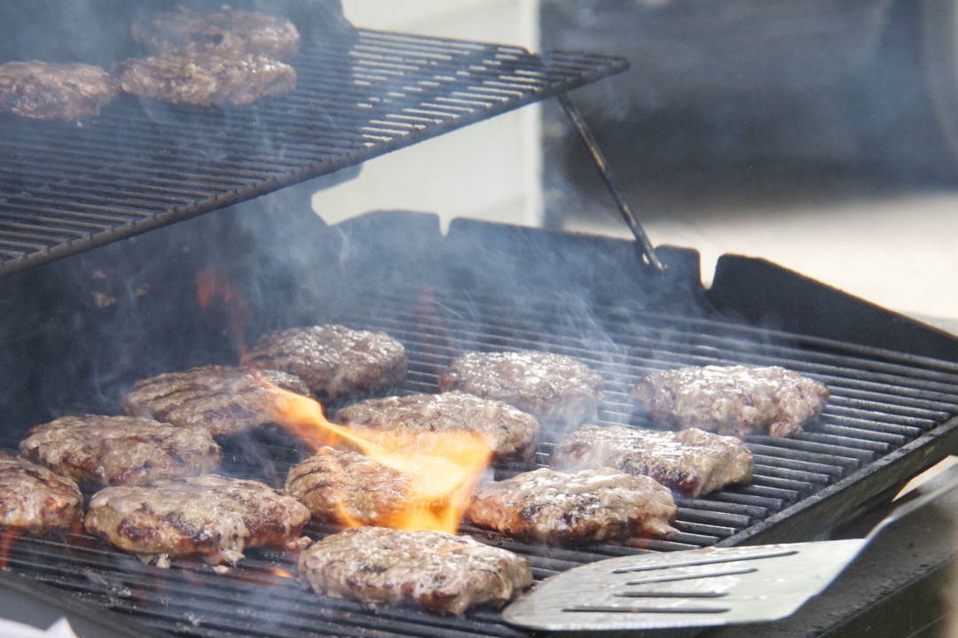 A slotted stainless steel grill spatula resting on a gas grill grate beside burgers over a flare-up