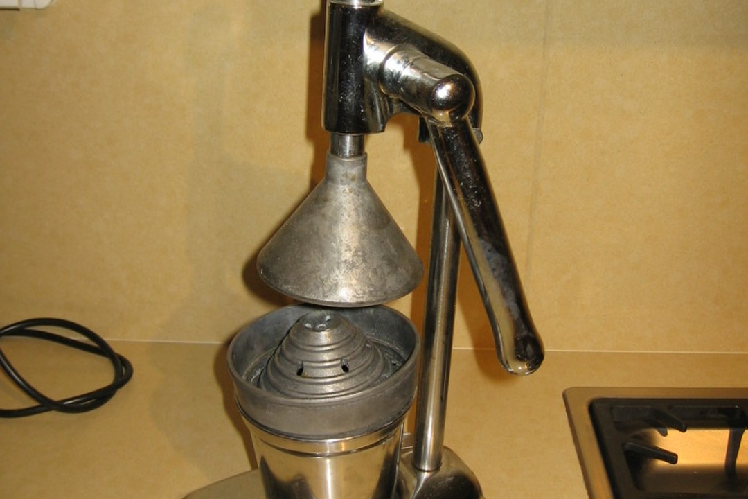 A metal lever-arm hand press juicer on a kitchen counter with the cone raised above the strainer cup
