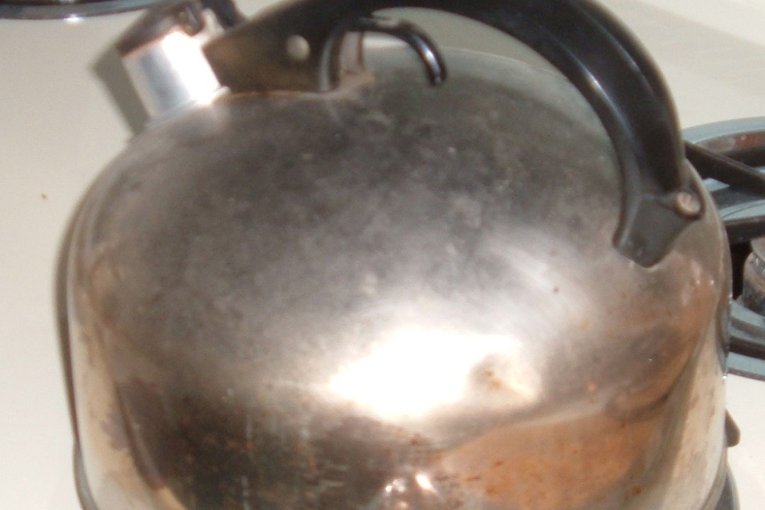 A stainless steel stovetop whistling kettle on a gas burner with heat discolouration and scorch staining across its body