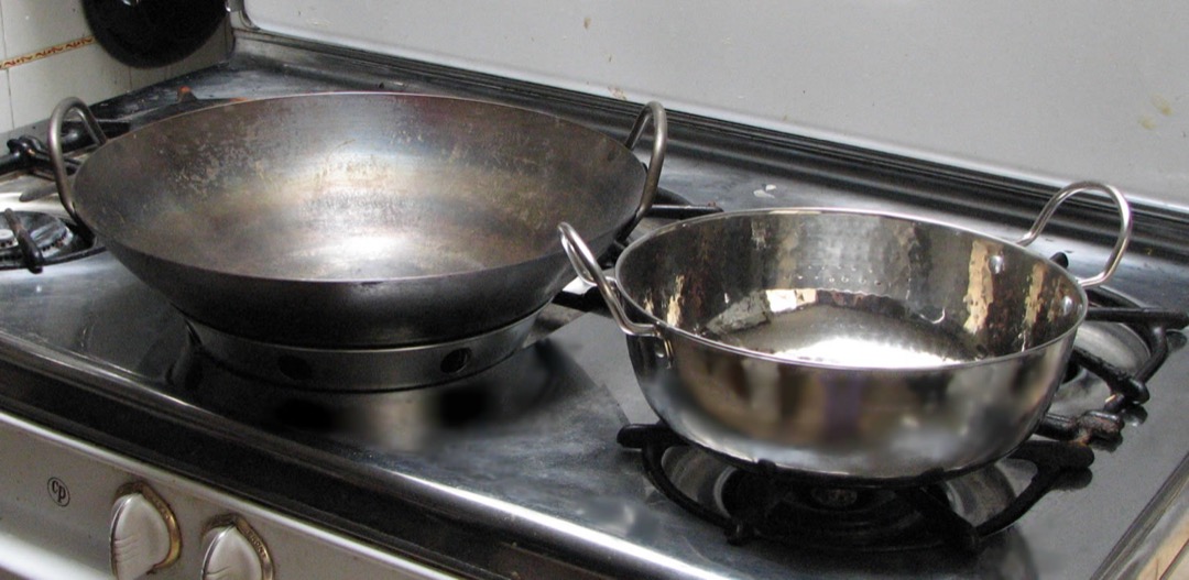 A used carbon steel wok with a patchy dark patina sitting on a gas stove next to a stainless steel karahi