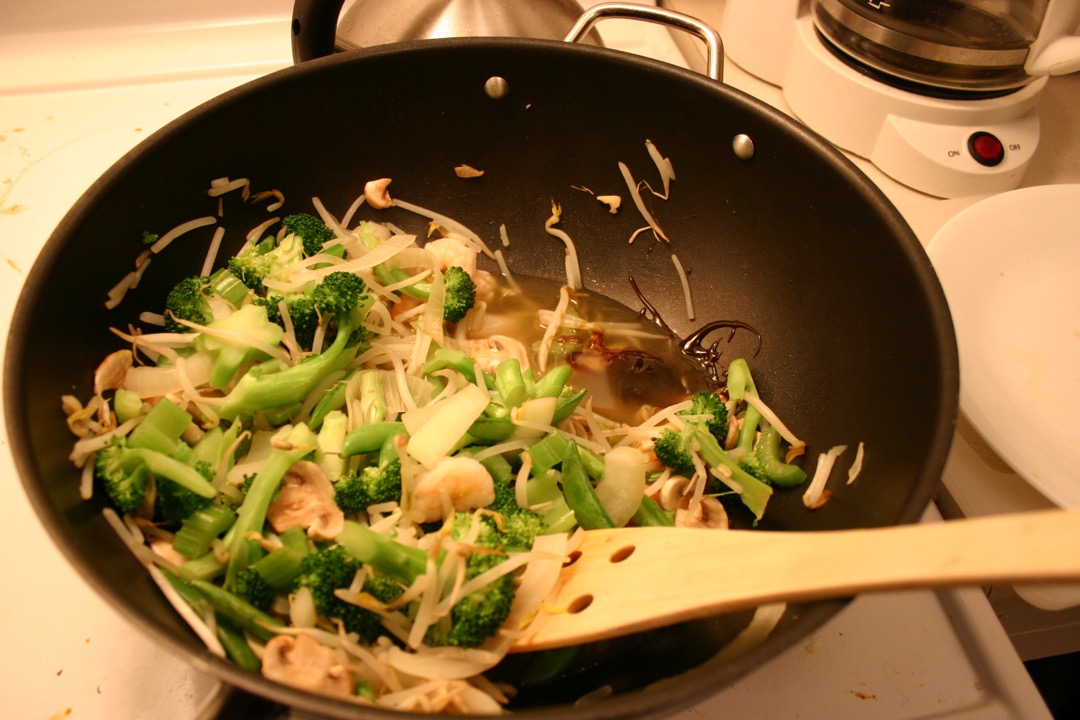 A dark wok on a home kitchen range part way through a vegetable stir-fry, with broccoli, snap peas, mushrooms and bean sprouts in the bowl and a wooden spatula laid across the base