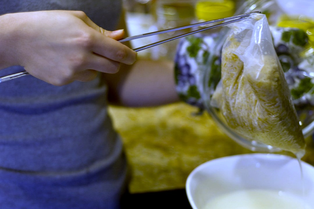 Long slim stainless steel kitchen tweezer tongs held in one hand, gripping a straining bag over a bowl
