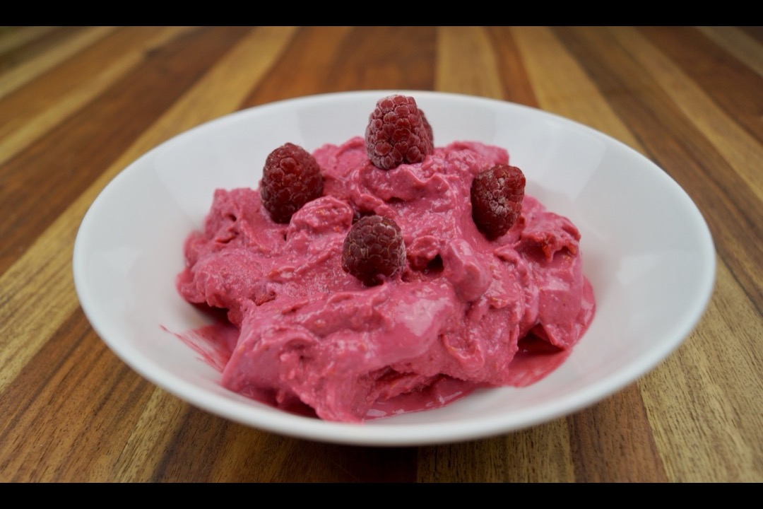 A white bowl of thick pink raspberry ice cream topped with whole raspberries, on a wooden counter