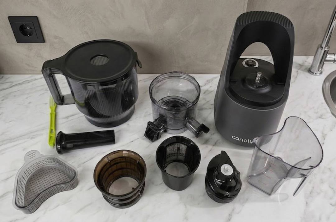 A masticating slow juicer fully disassembled on a white marble countertop, with the motor base, auger, mesh strainer basket, juicing chamber, pulp container, juice pitcher, feed-tube pusher and cleaning brush laid out in two rows